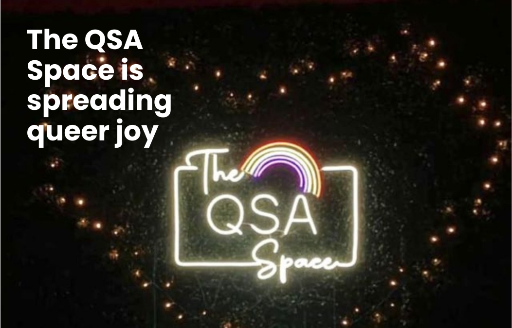 A neon sign with a rainbow and text - the QSA Space. Text on image - The QSA Space is spreading queer joy.