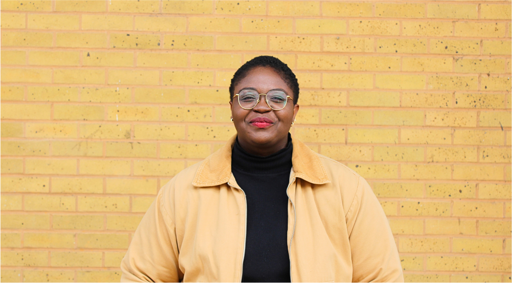 Fola Afolabi wearing glasses and a yellow jacket and a yellow jacket looking into the camera.