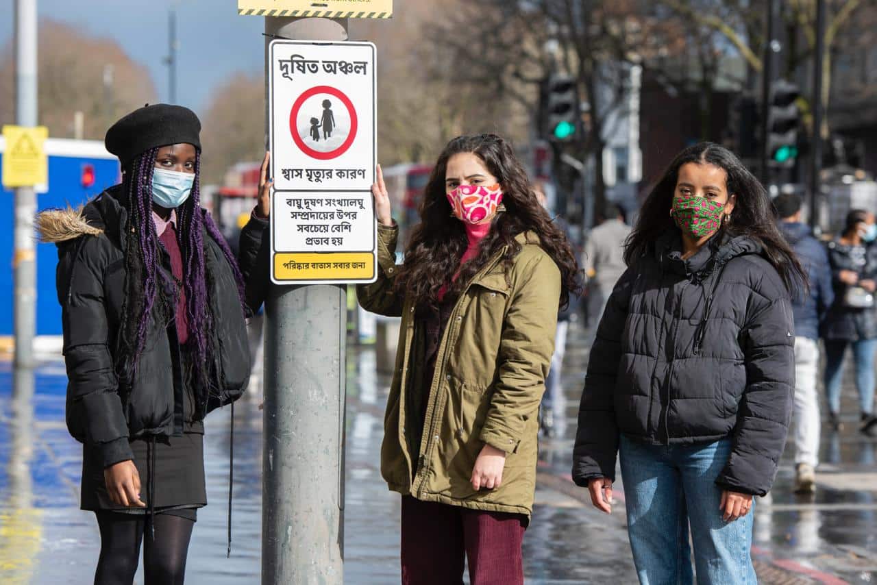 Three people standing by a 'pollution zone' sign wearing masks