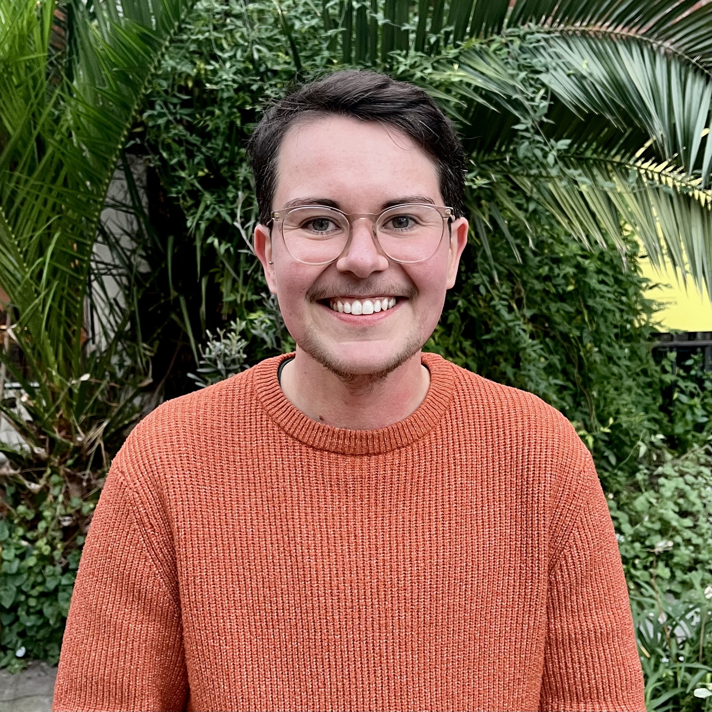 A person in an orange jumper wearing glasses and smiling for a camera